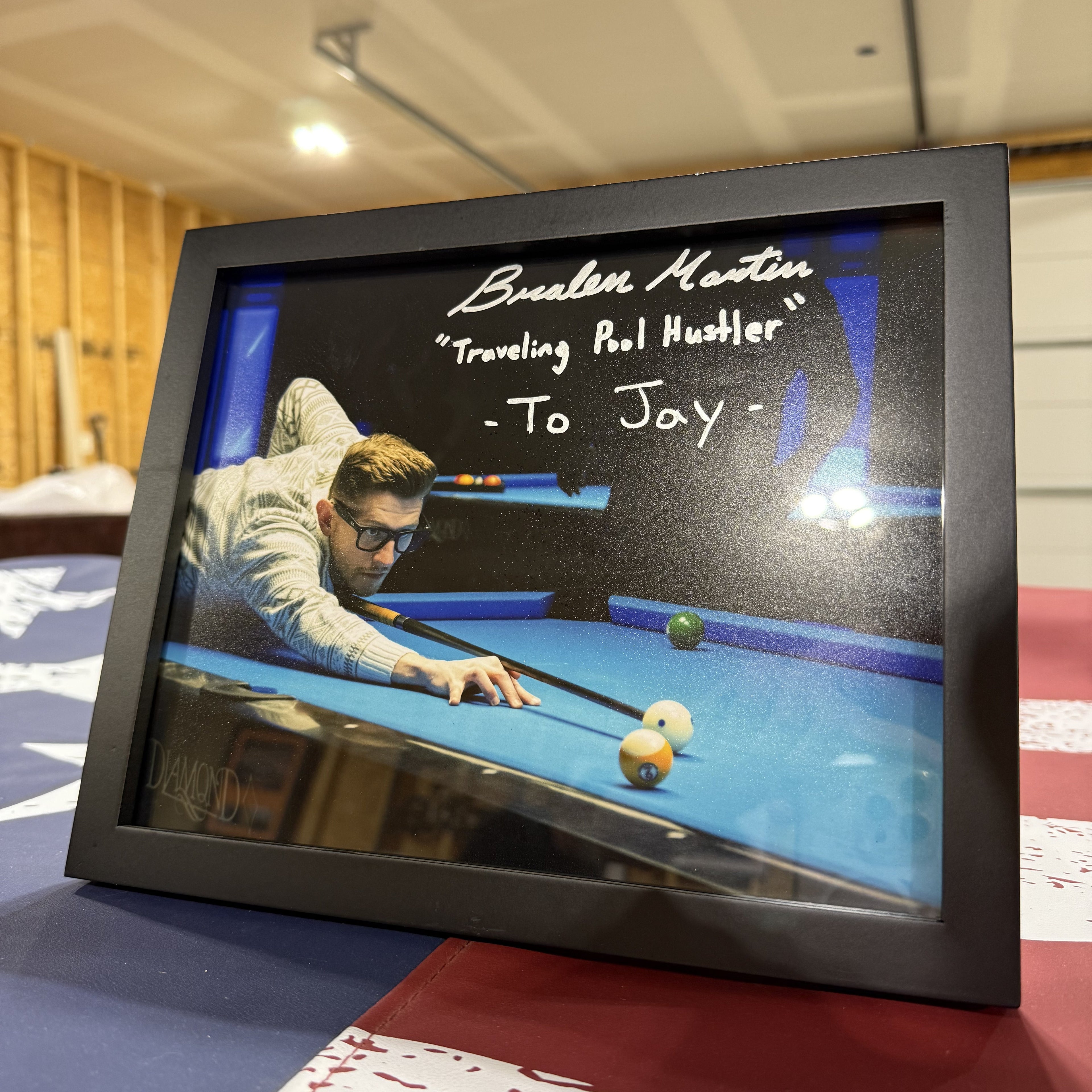 A black-framed custom photo print displayed on a red, white, and blue flag in a room. The photo inside the frame shows a man playing pool with a blue felt table, and the text 'Bralen Martin "Traveling Pool Hustler" -To Jay-' is written in silver script on the glass.